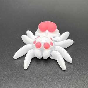May include: A white 3D printed spider with pink eyes and a pink body. The spider has eight legs and is sitting on a black surface.