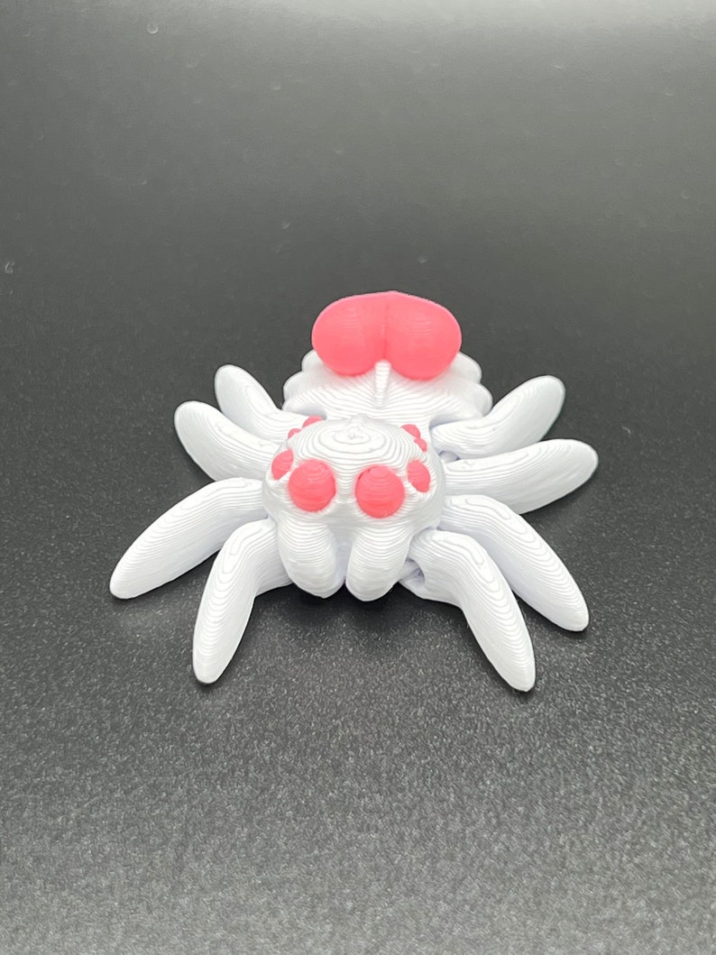 May include: A white 3D printed spider with pink eyes and a pink body. The spider has eight legs and is sitting on a black surface.