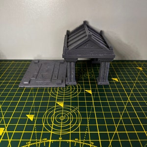 May include: A grey 3D printed model of a small building with a flat roof and a rectangular base. The building has a series of columns and a decorative roof.