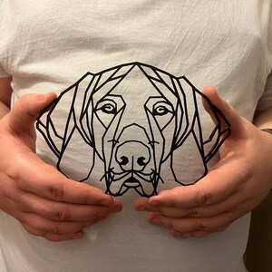 May include: A black geometric line art design of a dog's head, with a simple, modern style. The design is held in front of a white t-shirt.