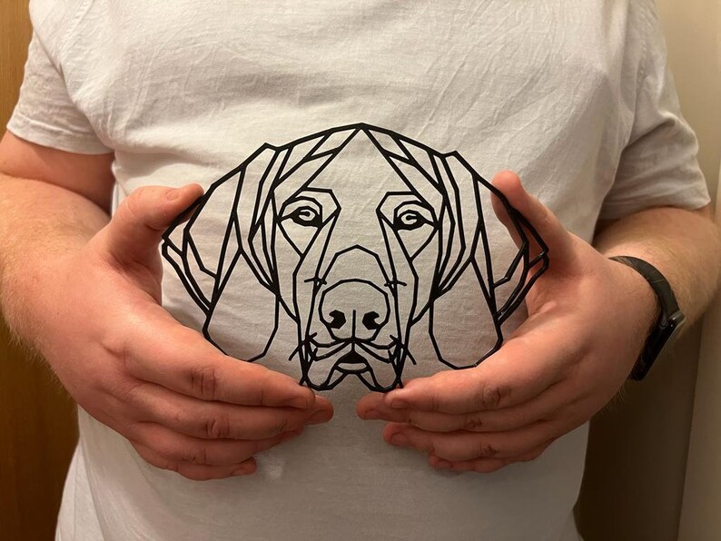 May include: A black geometric line art design of a dog's head, with a simple, modern style. The design is held in front of a white t-shirt.