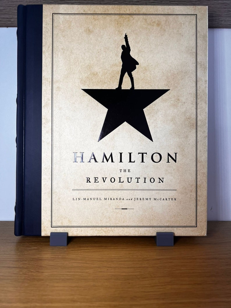 May include: A black and white book with the title "Hamilton: The Revolution" by Lin-Manuel Miranda and Jeremy McCarter. The cover features a silhouette of a person standing on a black star.