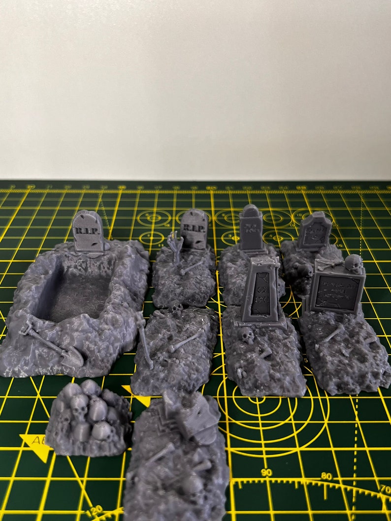 May include: A set of 3D printed miniature graveyard scenery pieces. The set includes a variety of tombstones, a grave, and piles of skulls. The tombstones are grey and have the inscription "R.I.P." on them. The grave is a rectangular hole in the ground with a shovel leaning against it. The skulls are piled up in a heap.