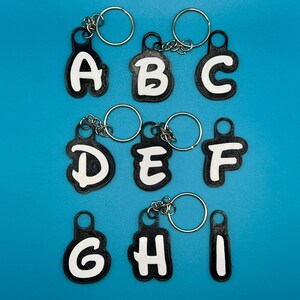 Op de afbeelding: Negen zwarte sleutelhangers met witte letters die het alfabet van A tot en met I spellen. Elke sleutelhanger heeft een zilveren ring.