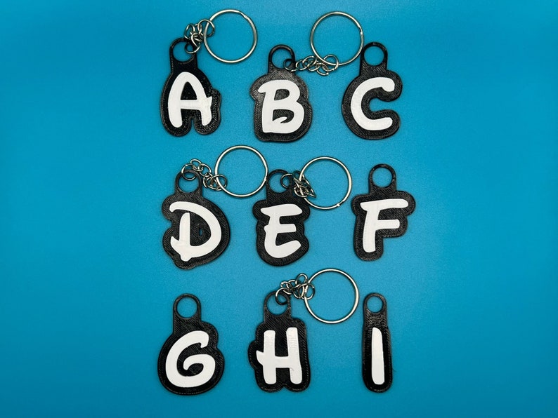 Op de afbeelding: Negen zwarte sleutelhangers met witte letters die het alfabet van A tot en met I spellen. Elke sleutelhanger heeft een zilveren ring.