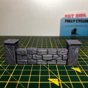 May include: A miniature stone wall with two pillars, made of gray plastic. The wall is textured to look like rough stone.