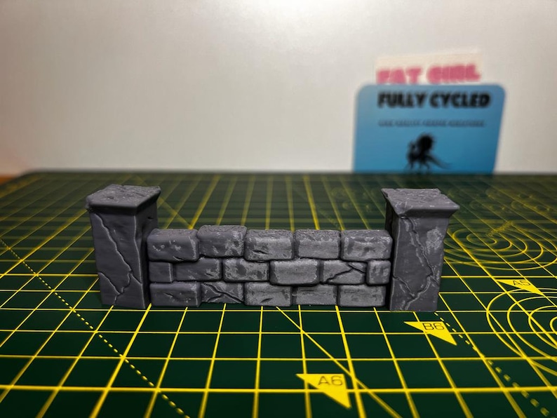 May include: A miniature stone wall with two pillars, made of gray plastic. The wall is textured to look like rough stone.