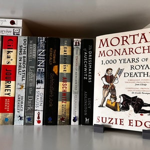 May include: A bookshelf with a variety of books, including "The Tattooist of Auschwitz" and "Mortal Monarchs: 1,000 Years of Royal Deaths" by Susie Edge. The book "Mortal Monarchs" is on a stand in front of the bookshelf.