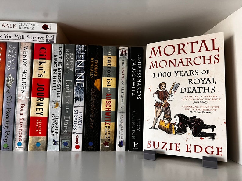 May include: A bookshelf with a variety of books, including "The Tattooist of Auschwitz" and "Mortal Monarchs: 1,000 Years of Royal Deaths" by Susie Edge. The book "Mortal Monarchs" is on a stand in front of the bookshelf.