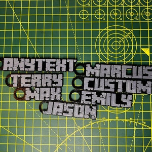 May include: A close-up of a green cutting mat with several personalized name tags. The tags are made of a gray material with black lettering. The names on the tags are "Anytext", "Marcus", "Terry", "Custom", "Max", "Emily", and "Jason".