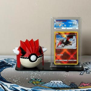 Könnte beinhalten: Eine rot-weiße Pokémon-Figur und eine Radiant Charizard-Sammelkarte in einer Schutzhülle. Die Karte ist als Gem Mint 10 bewertet. Die Karte zeigt eine feurige Drachenillustration.