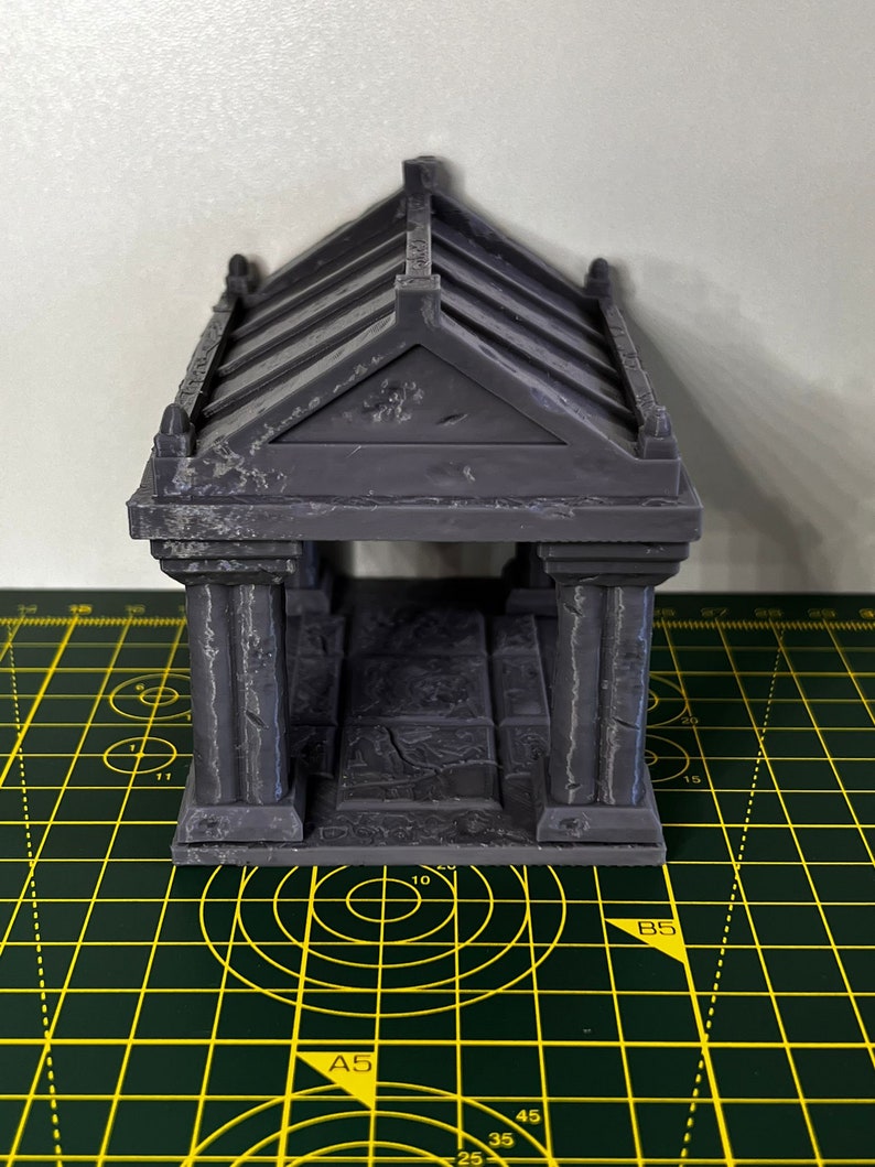 May include: A gray 3D printed miniature building with a gabled roof and columns. The building is sitting on a green cutting mat.