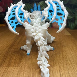 Puede incluir: Esqueleto de drag&oacute;n de pl&aacute;stico blanco con alas azules. El drag&oacute;n est&aacute; de pie sobre una superficie de madera con las alas extendidas.
