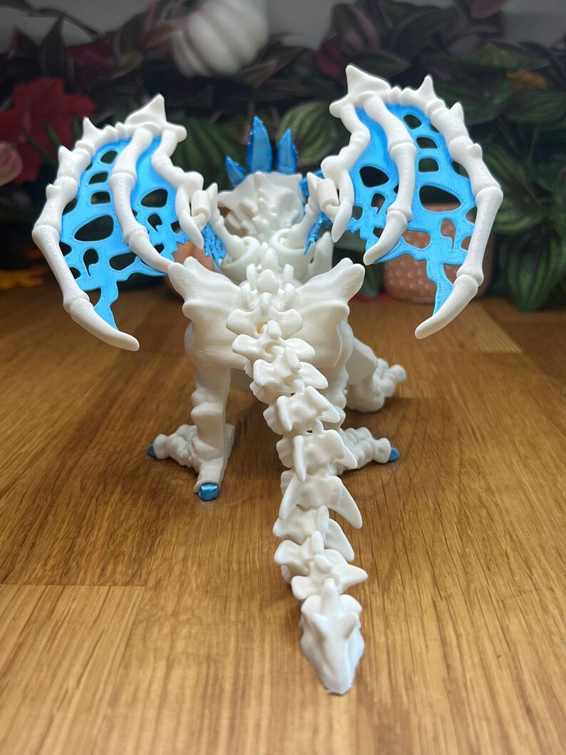 Puede incluir: Esqueleto de drag&oacute;n de pl&aacute;stico blanco con alas azules. El drag&oacute;n est&aacute; de pie sobre una superficie de madera con las alas extendidas.