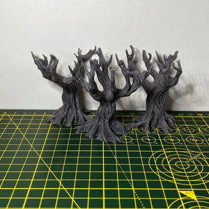 May include: Three grey, 3D printed, miniature, skeletal trees with gnarled branches and roots. The trees are arranged in a cluster on a green cutting mat.