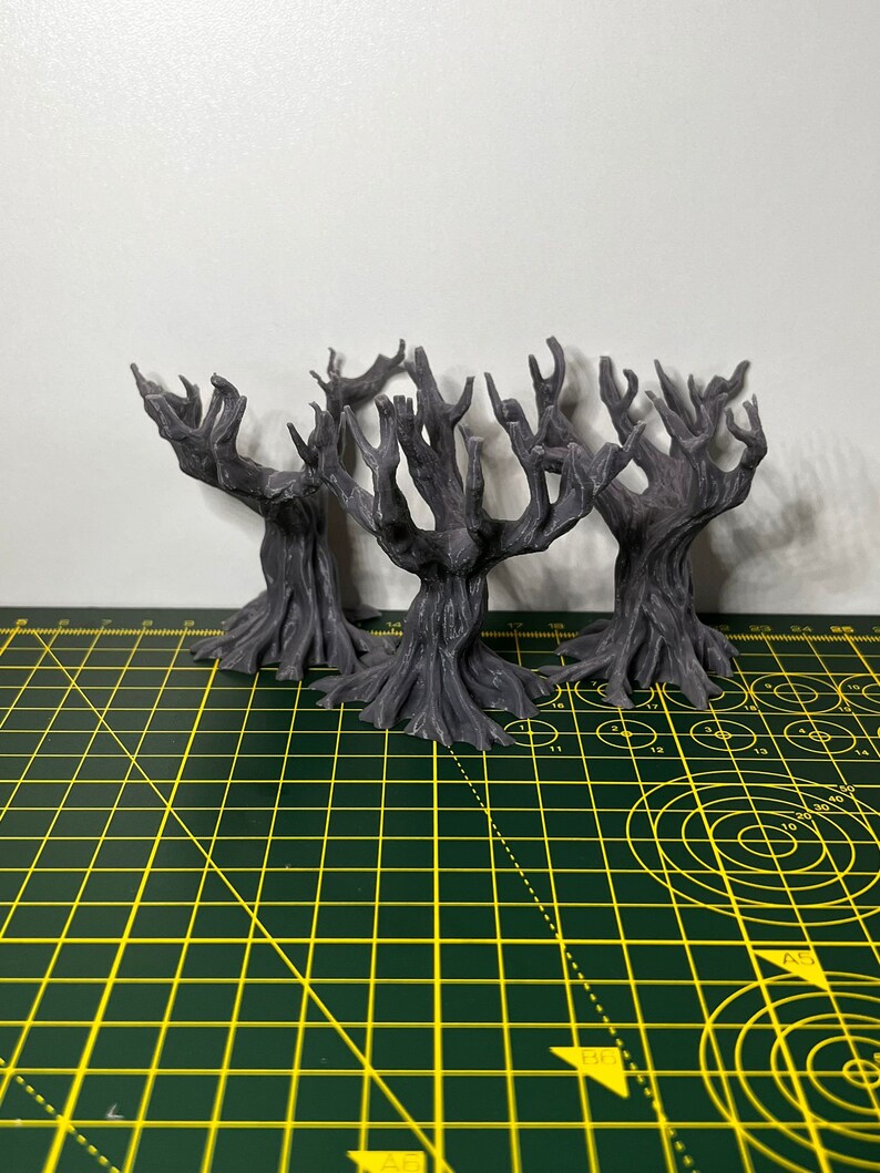 May include: Three grey, 3D printed, miniature, skeletal trees with gnarled branches and roots. The trees are arranged in a cluster on a green cutting mat.