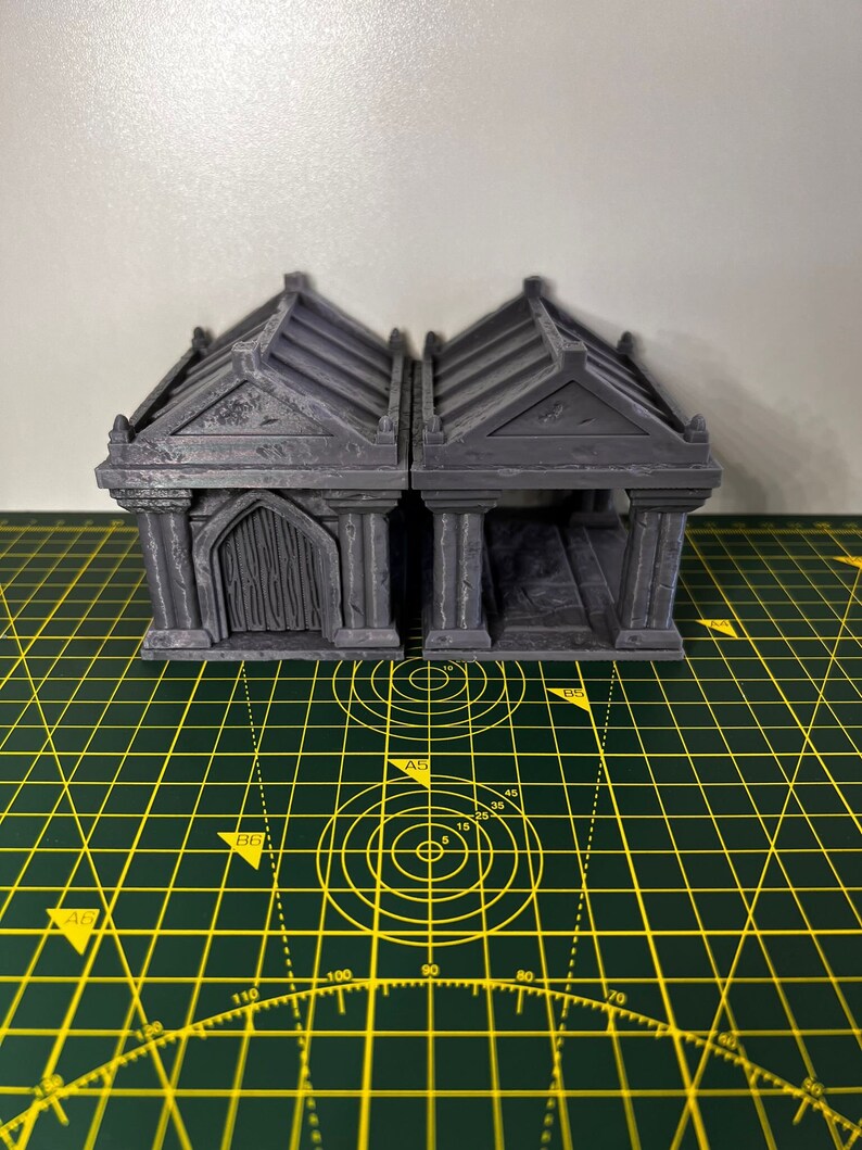 May include: Two grey, miniature, 3D printed mausoleums with arched doorways and detailed stonework. The mausoleums are sitting on a green cutting mat with yellow markings.