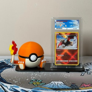 Op de afbeelding: Een oranje en witte Pokémon-balfiguur met een vlamaccessoire naast een beoordeelde Radiant Charizard-ruilkaart. De kaart zit in een beschermhoes en heeft een Charizard-illustratie. De kaart is beoordeeld als 10 GEM MINT.