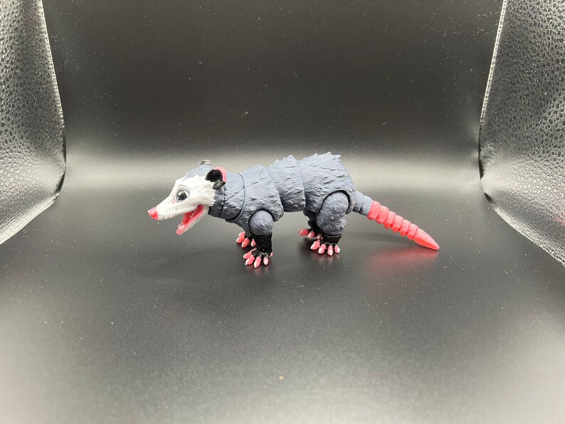 May include: A black and white toy opossum with a pink nose and tail. The toy has articulated limbs and a textured body.