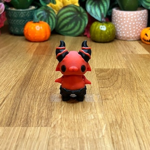 Puede incluir: Una pequeña figura de diablo roja y negra con cuernos y cola. La figura está de pie sobre una superficie de madera.