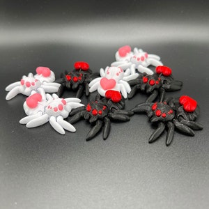 May include: A group of eight small, 3D printed spiders with red hearts on their abdomens.  Half of the spiders are black and half are white.