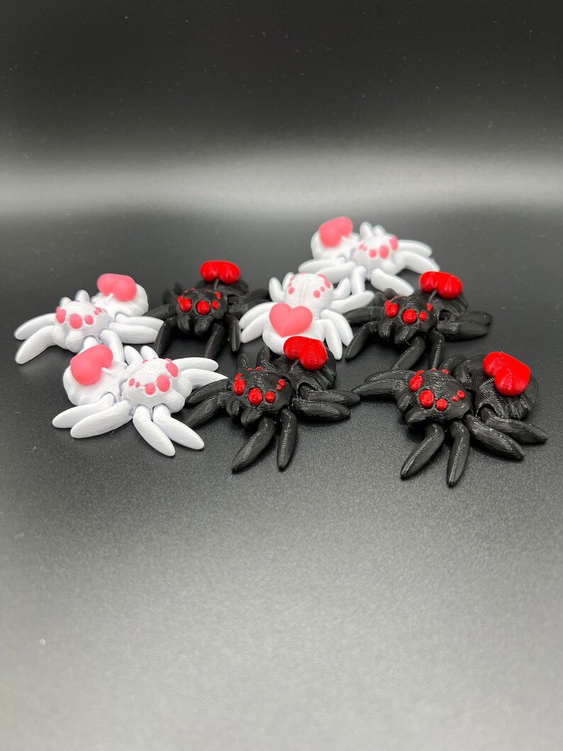 May include: A group of eight small, 3D printed spiders with red hearts on their abdomens.  Half of the spiders are black and half are white.