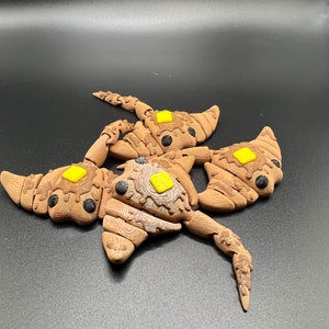 May include: A brown and black ceramic sculpture of a creature with three heads and yellow accents. The sculpture is made to look like a pastry with a brown glaze and yellow icing.