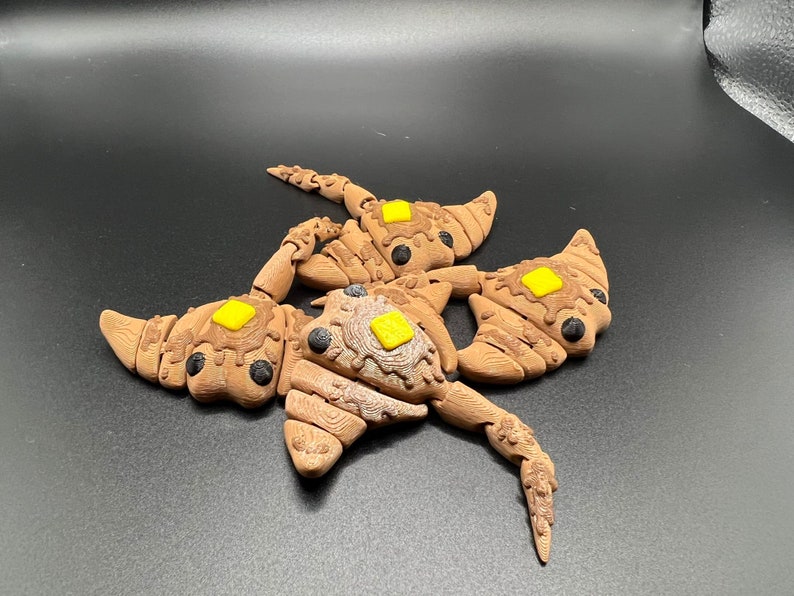 May include: A brown and black ceramic sculpture of a creature with three heads and yellow accents. The sculpture is made to look like a pastry with a brown glaze and yellow icing.