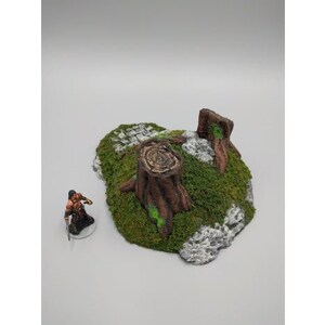 May include: A miniature tabletop gaming terrain piece featuring a tree stump with green moss and a rocky base.