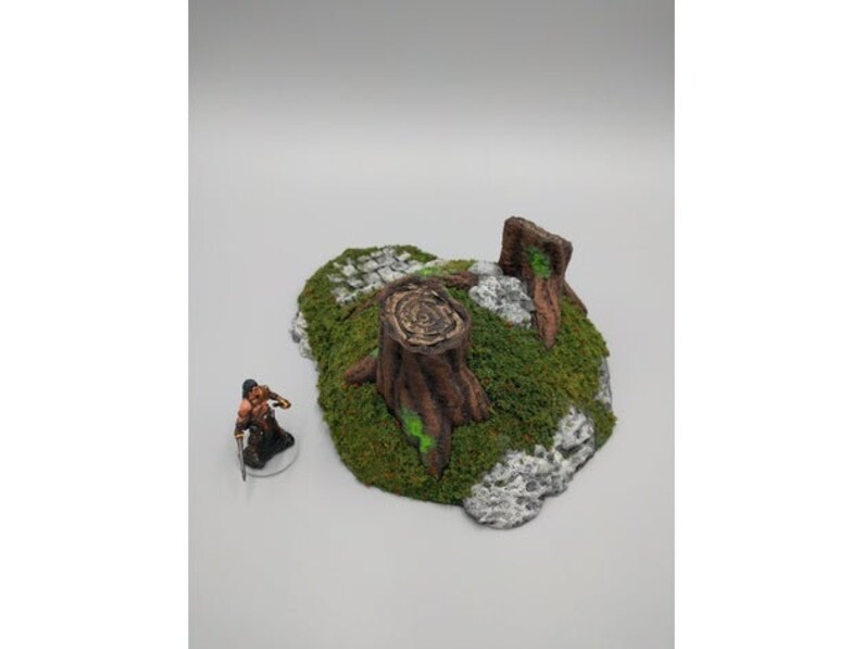 May include: A miniature tabletop gaming terrain piece featuring a tree stump with green moss and a rocky base.