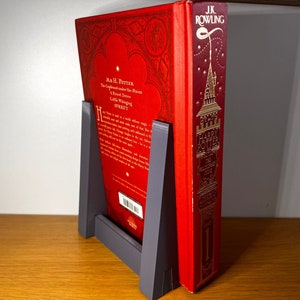 May include: A red hardcover book with gold lettering titled "Harry Potter and the Philosopher's Stone" by J.K. Rowling. The book is propped up by a gray bookend.