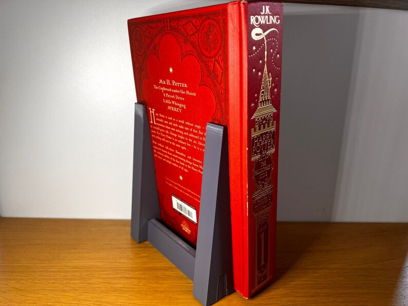 May include: A red hardcover book with gold lettering titled "Harry Potter and the Philosopher's Stone" by J.K. Rowling. The book is propped up by a gray bookend.