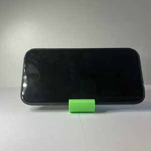 May include: A black smartphone is propped up on a bright green phone stand. The phone is on a white surface.