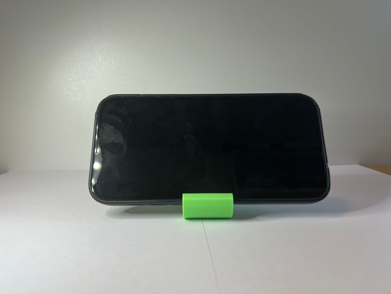 May include: A black smartphone is propped up on a bright green phone stand. The phone is on a white surface.