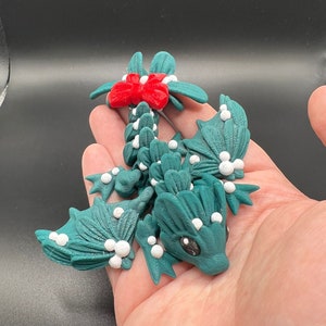 May include: A green dragon figurine with white and red accents. The dragon is decorated with white dots and has a red bow on its tail. The dragon is made of polymer clay and is a Christmas-themed decoration.