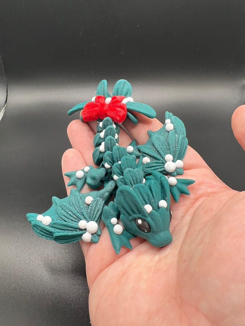 May include: A green dragon figurine with white and red accents. The dragon is decorated with white dots and has a red bow on its tail. The dragon is made of polymer clay and is a Christmas-themed decoration.