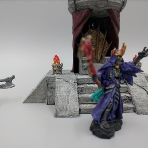 May include: A miniature figure of a skeletal figure wearing a purple robe and a crown, holding a staff. The figure is standing on a stone platform with steps leading up to a throne with a red curtain.