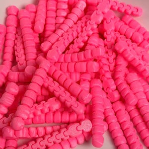 Puede incluir: Un montón de conectores de plástico rosa brillante segmentados. Cada pieza tiene una serie de segmentos redondeados conectados en forma de cadena. Los conectores están diseñados para entrelazarse, creando una estructura flexible. La imagen está bien iluminada, resaltando el color y la textura vibrantes.