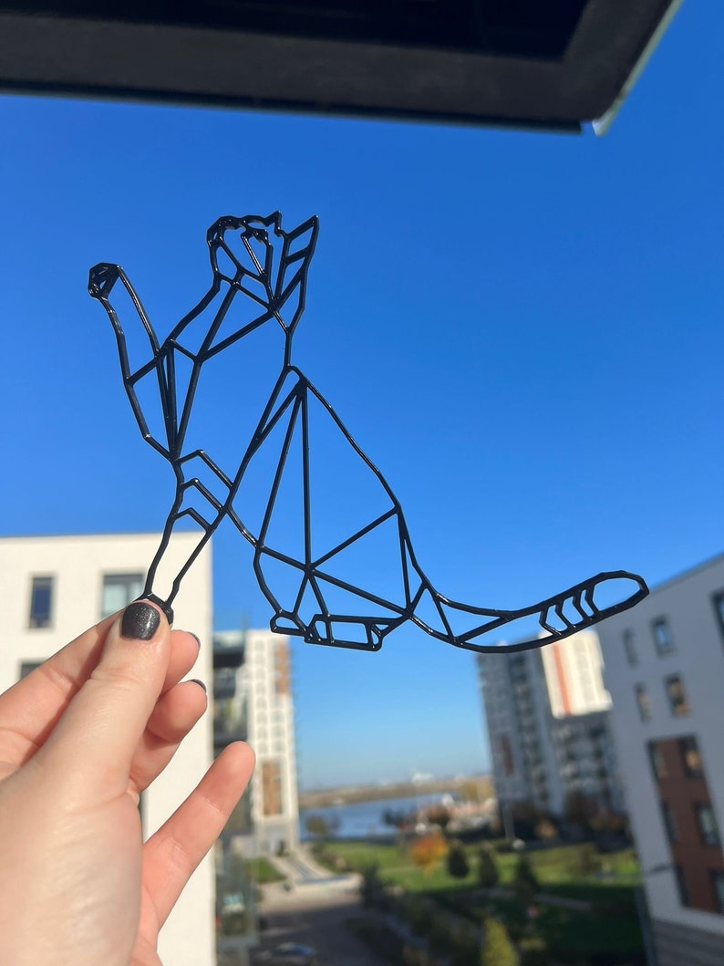 May include: A black geometric cat silhouette, made of wire, is held in front of a window with a view of buildings and a river.