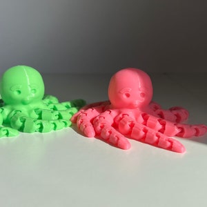 以下が含まれることがあります： 顔と8本の関節のある触手が詳細に描かれた、緑とピンクの2つの3Dプリントされたタコの人形です。