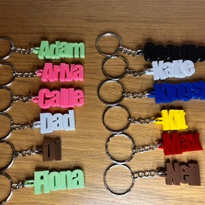 Op de afbeelding: Een verzameling van 10 gepersonaliseerde sleutelhangers met namen in verschillende kleuren. De namen zijn Adam, Ariya, Callie, Dad, Di, Fiona, George, Katie, Kingsley, Kit, Max en Neil.