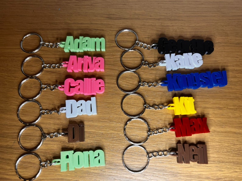 Op de afbeelding: Een verzameling van 10 gepersonaliseerde sleutelhangers met namen in verschillende kleuren. De namen zijn Adam, Ariya, Callie, Dad, Di, Fiona, George, Katie, Kingsley, Kit, Max en Neil.