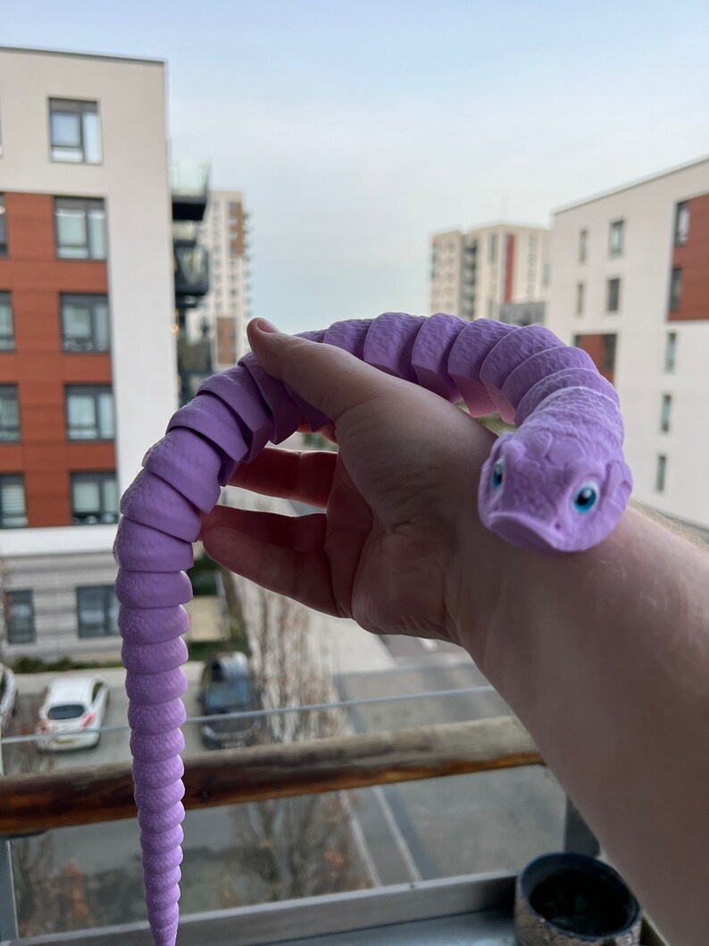 May include: A purple segmented toy snake with a textured surface and large blue eyes. The snake is coiled in a person's hand.