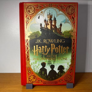 May include: A red hardcover book with a gold-embossed design. The book features an illustration of a castle with a blue sky and green trees. The title "J.K. Rowling Harry Potter and the Philosopher's Stone" is printed in gold lettering on the front cover.
