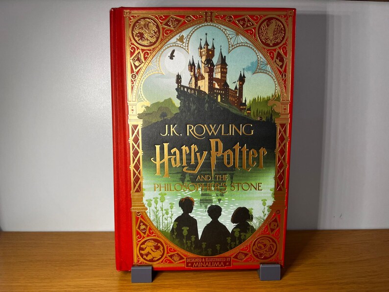 May include: A red hardcover book with a gold-embossed design. The book features an illustration of a castle with a blue sky and green trees. The title "J.K. Rowling Harry Potter and the Philosopher's Stone" is printed in gold lettering on the front cover.