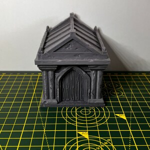 May include: A gray, 3D printed miniature building with a gabled roof and a door. The building has a textured surface and is sitting on a green cutting mat.