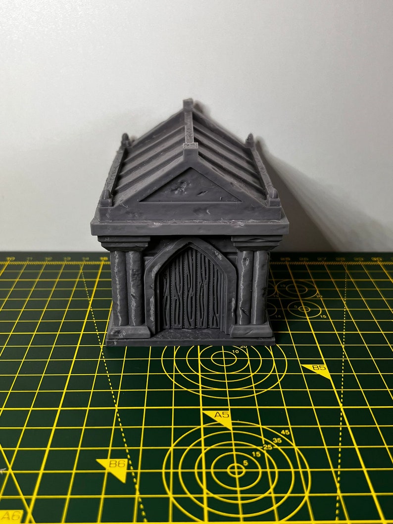May include: A gray, 3D printed miniature building with a gabled roof and a door. The building has a textured surface and is sitting on a green cutting mat.