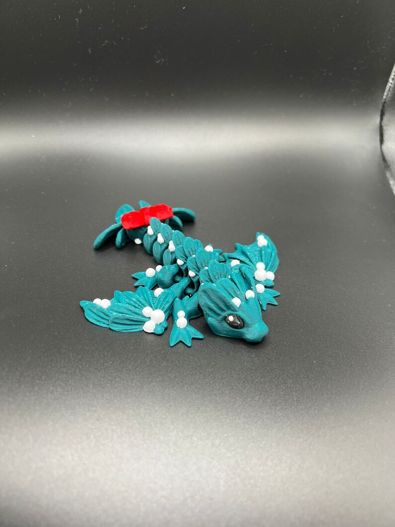 May include: Teal blue 3D printed dragon figurine with white accents and a red stripe on its back. The dragon has a small, detailed face with black eyes.
