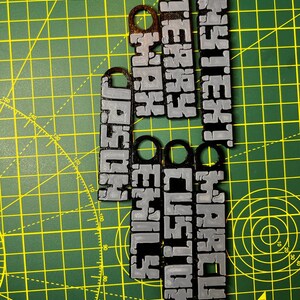 May include: Five black and silver keychains with different names printed on them. The names are "ANYTEXT", "MARCUS", "TERRY", "CUSTOM", "MAX", "EMILY", and "HOSHO".