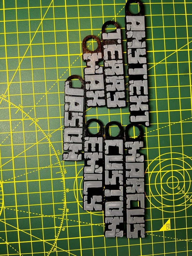 May include: Five black and silver keychains with different names printed on them. The names are "ANYTEXT", "MARCUS", "TERRY", "CUSTOM", "MAX", "EMILY", and "HOSHO".